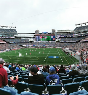 A large sports stadium filled with spectators. The field is green with white markings, and players in red and white uniforms are actively engaged in a game. The stands are packed with people wearing various colored clothing, and some sections of the crowd are more densely populated. Digital screens and advertisements are visible around the stadium, adding to the vibrant atmosphere.