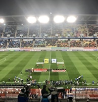 A large soccer stadium with bright overhead lights and a well-maintained green field. Teams are lined up in the center alongside officials, and there is a group gathered nearby, possibly for a pre-match ceremony. The stadium is filled with spectators wearing colorful clothing, and banners and advertisements are visible along the edges of the field. Cameras and officials are positioned on the sidelines to capture the event.