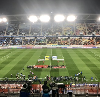 A large soccer stadium with bright overhead lights and a well-maintained green field. Teams are lined up in the center alongside officials, and there is a group gathered nearby, possibly for a pre-match ceremony. The stadium is filled with spectators wearing colorful clothing, and banners and advertisements are visible along the edges of the field. Cameras and officials are positioned on the sidelines to capture the event.