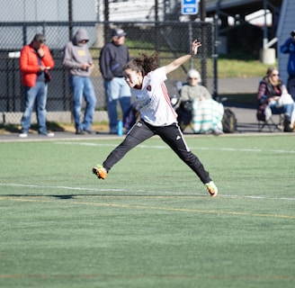 A person is mid-air on a sports field, appearing to be involved in an athletic activity. Several spectators are on the sidelines, some seated and others standing, with one individual capturing the event with a camera. The background includes a fence and various casual observers, some of whom are wearing winter clothing.
