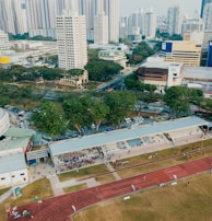 An aerial view of an urban area featuring a sports complex with a track field and a grandstand. Surrounding the sports area are numerous high-rise buildings, greenery, and roads with vehicles. The image captures people engaging in activities on the field.