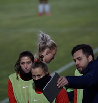 A group of individuals on a sports field are wearing green vests over red attire. One person is holding a tablet and seems to be discussing or showing something to the others. The atmosphere suggests a sports event, likely soccer, indicated by their clothing and the setting. The background is a grassy field.