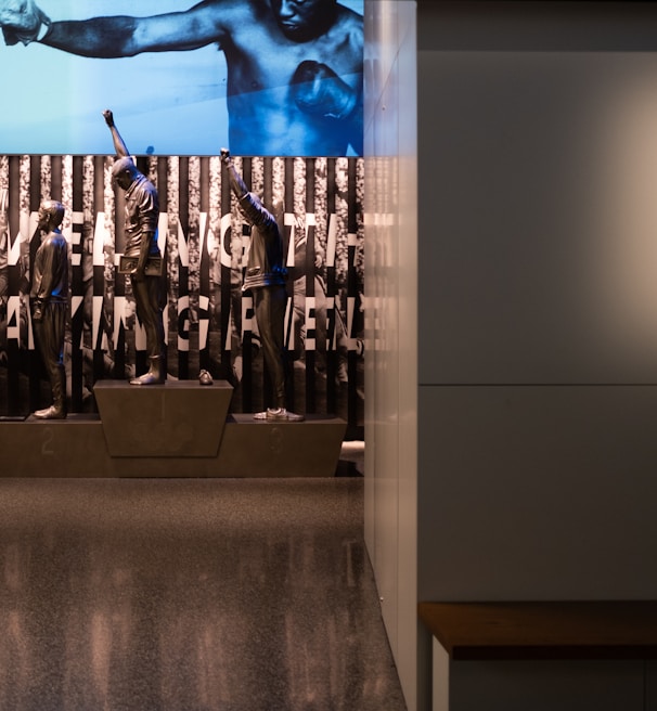 A dimly lit display features statues of athletes standing on a podium. They are positioned in front of a backdrop with the words 'LEVELING THE PLAYING FIELD' in bold letters. On the wall to the right, a softly lit quote is partially visible. The overall ambiance suggests a museum or exhibition setting.