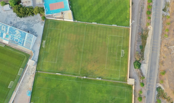 An aerial view of a sports complex featuring multiple green grass fields, including football pitches with marked goalposts. Adjacent to the fields is a blue-roofed building and a smaller area with a tennis or basketball court. Surrounding the complex, there's a road lined with trees and a dirt area with sparse vegetation.