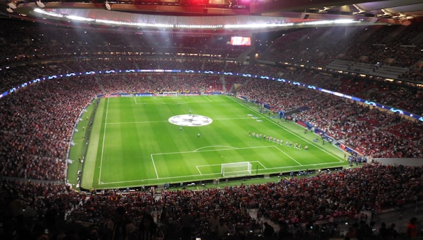 A large, packed stadium with bright lights illuminates a green soccer field. The scene is vibrant with a massive audience surrounding the pitch, visibly excited and engaged. The center of the field displays a prominent circular logo, suggesting a major sporting event. Players and staff can be seen on the field, likely preparing for a match.