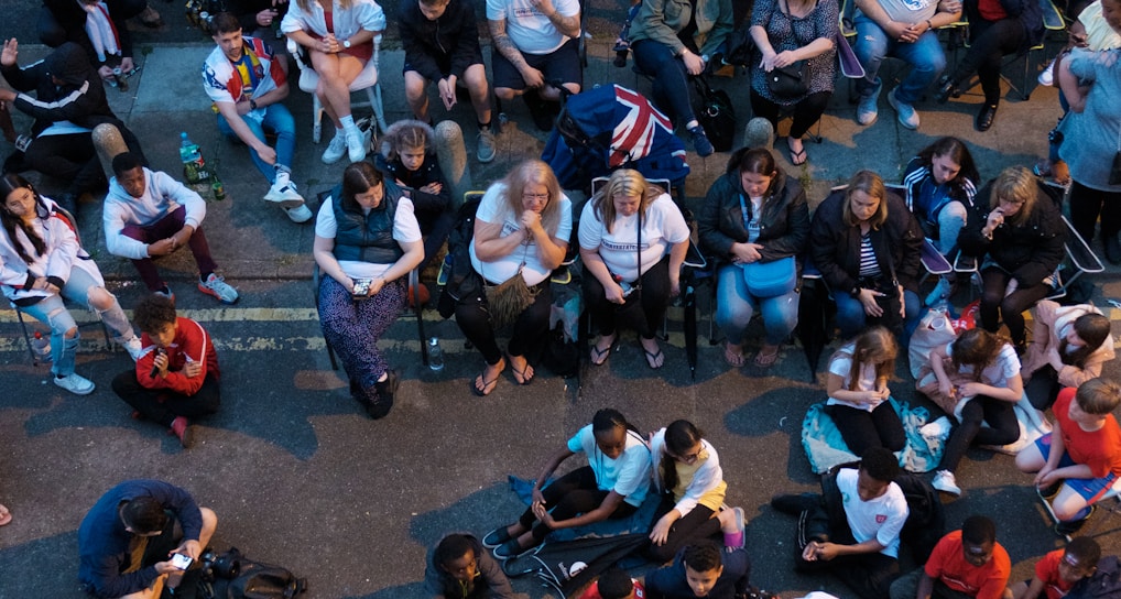 A large group of people gathered outdoors, watching a screen that is displaying a man. The crowd is diverse in age and appears interested and engaged, with many sitting closely together, some standing. Many are dressed casually, some in sports attire, and a few waving or holding flags. The event seems to have a communal, festive atmosphere.