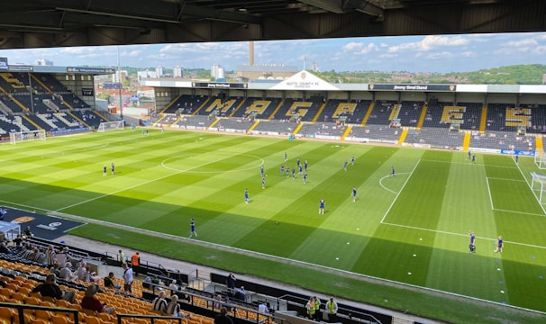A sports stadium with a well-maintained grass field is depicted. Players are spread across the field, engaging in warm-up activities. The stands are partially filled with spectators, and the stadium is surrounded by urban buildings under a partly cloudy sky.