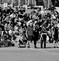A large crowd of people is gathered, with some standing and others sitting on bleachers. Several individuals are wearing sports attire, and there is a field visible in the foreground with a marker indicating 50 meters. The scene appears to be a sporting event, with a diverse group of spectators.