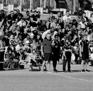 A large crowd of people is gathered, with some standing and others sitting on bleachers. Several individuals are wearing sports attire, and there is a field visible in the foreground with a marker indicating 50 meters. The scene appears to be a sporting event, with a diverse group of spectators.