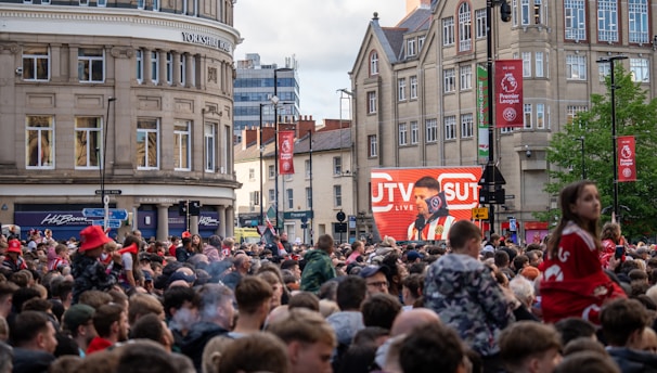 A large crowd of people, many wearing red and white, gather in a city square. They are watching an event broadcast on a large screen, which displays a man speaking into a microphone with logos suggesting a sporting event. Surrounding buildings are historical and commercial, with banners indicating a connection to the Premier League.