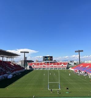 A large sports stadium with red and blue seating overlooks a green field with a goalpost at the center. A few people are scattered across the field, with some tents and a gathering of individuals on the right side. The sky is clear and blue with some clouds visible in the distance. A large screen or scoreboard is situated at the far end of the field.
